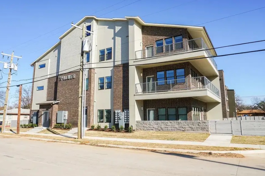 Modern three-story apartment building with large balconies, brick and light siding exterior, and a sidewalk along the street in front. Power lines and a utility pole are visible in the foreground at The Palmer Fort Worth.