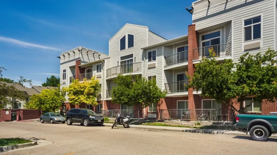 A modern three-story apartment building, The Social West® Fort Collins features balconies, brick and light siding exterior, trees lining the sidewalk, and several parked vehicles on a sunny day.