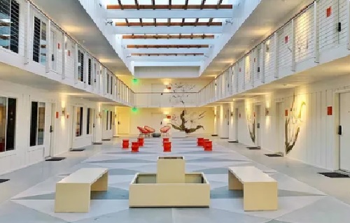 A bright, modern indoor space at The Mod at Riverwalk Reno features white walls, benches, and red chairs arranged in the center. Natural light streams through a skylight onto painted plants, creating a welcoming communal lounge area.