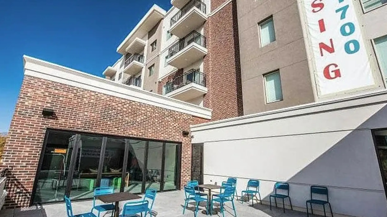 Outdoor patio area with blue chairs and tables in front of Towerview Apartments Ogden, a modern building with balconies; a large leasing banner hangs on the wall.