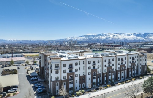 A modern, multi-story apartment building, The Republic Reno, with a white and brown exterior sits in an urban area, framed by snow-capped mountains and a city skyline visible in the background under a clear blue sky.