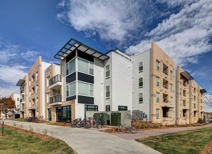 Modern, three-story apartment building with balconies, large windows, and a beige and white exterior at U Club on 28th Boulder. Several bicycles are parked outside on a sunny day with blue sky and clouds.