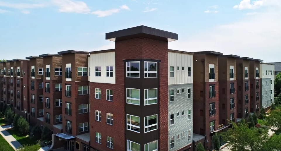 A modern multi-story apartment building, University House Denver blends red brick and light-colored siding, featuring large windows and landscaped greenery under a partly cloudy sky.