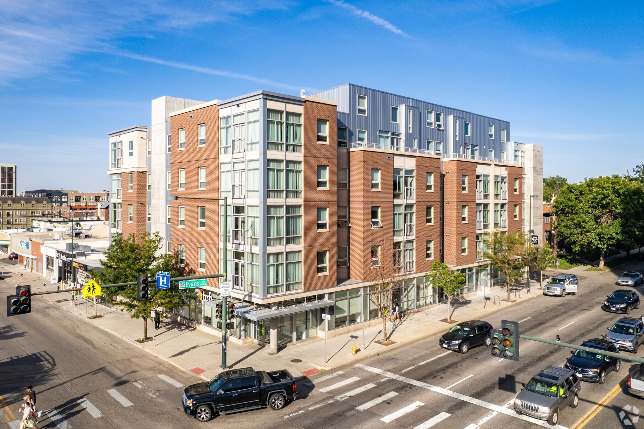 University Lofts, a modern five-story apartment building with a brick and glass exterior, sits on a busy city corner; cars, bikes, and pedestrians move through the intersection under a clear blue sky.