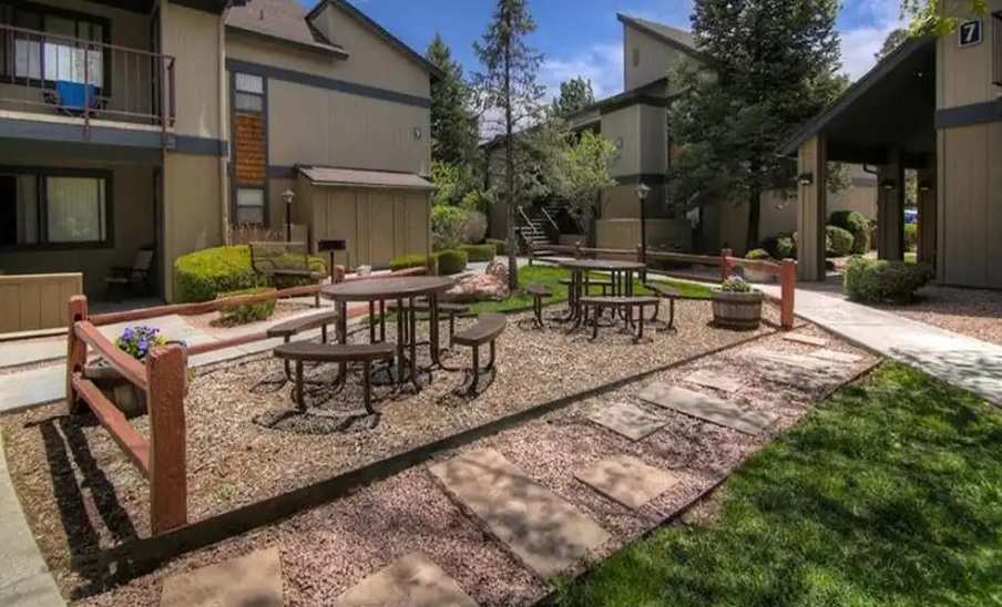 Outdoor apartment courtyard at University Square Apartments Flagstaff with picnic tables, benches, and a gravel area bordered by wooden rails. Surrounding buildings feature balconies, landscaped greenery, and walking paths with flat stone pavers.