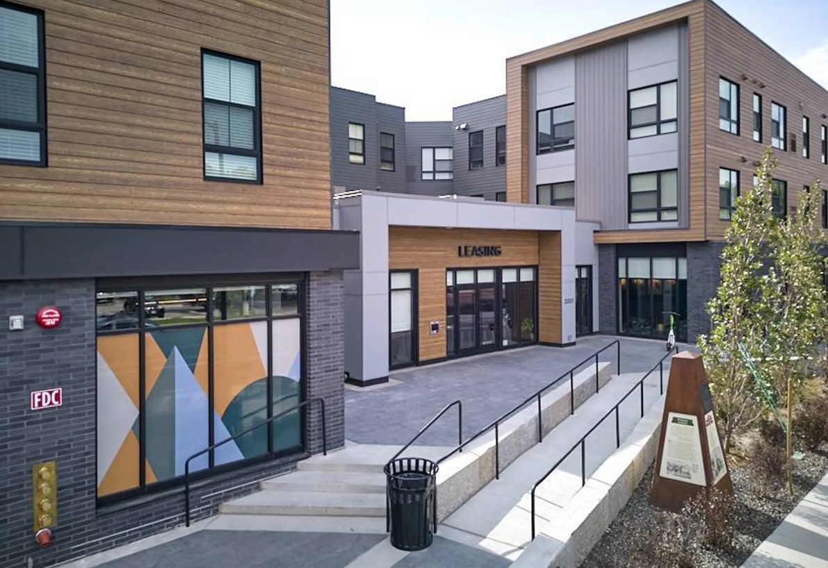 Modern apartment building, VERVE Boise, features wood and gray paneling, large windows, and a leasing office entrance. Colorful geometric art brightens a ground-floor window. A walkway with handrails and landscaping welcomes you in front.