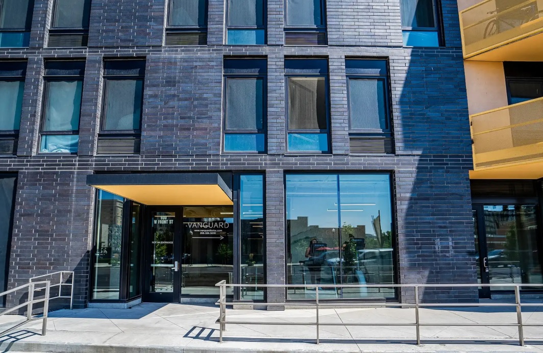 A modern building exterior at Vanguard Boise features dark brick, large windows, a sliding glass entrance, yellow awnings, a metal handrail, and a sidewalk out front. Reflections of trees and sky shimmer across the glass.
