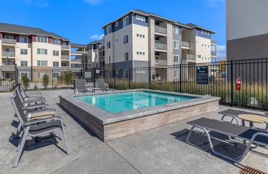 A small outdoor pool surrounded by lounge chairs and a black metal fence, set within the courtyard of Village East Apartments & Townhomes Boise under a clear blue sky.