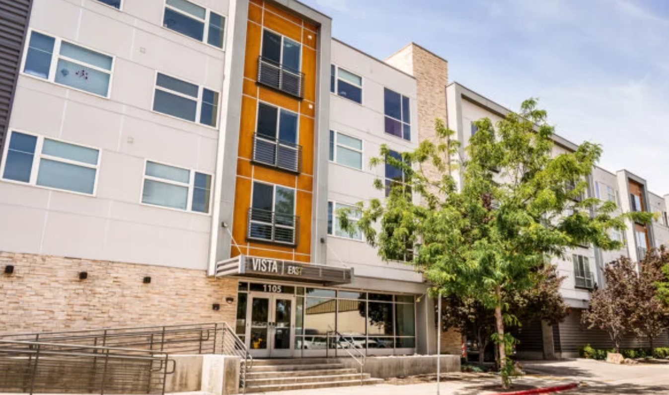 A modern four-story apartment building, The Vista Apartments Boise features large windows, a tree in front, and a sign above the entrance reading VISTA and 1105. The building boasts light-colored walls and stone accents.