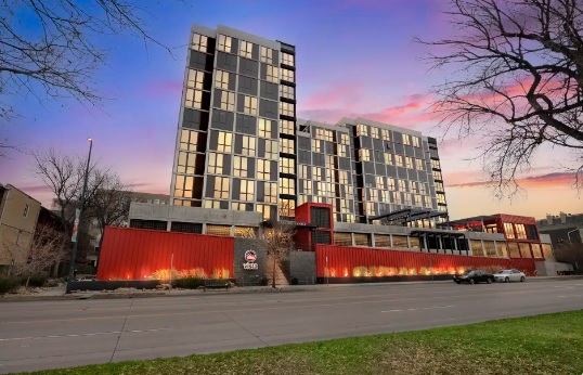 A modern multi-story building with large windows reflecting the colorful sunset sky creates a striking vista in Denver, Colorado. Red and gray exteriors, parked cars, and leafless trees frame the vibrant urban scene.