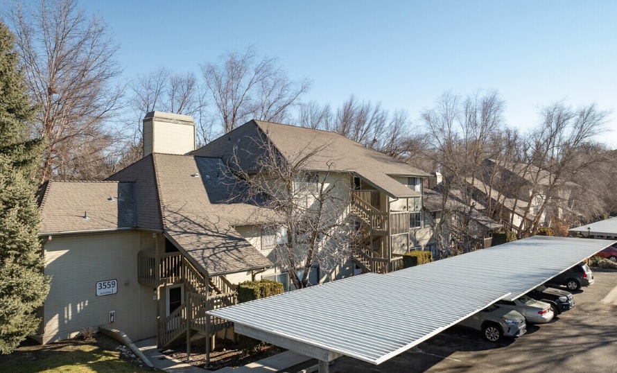 An apartment building with tan siding and a steep roof is shown in the Woodbine Boise area. A metal carport covers parked cars in front, surrounded by leafless trees under a clear blue sky. Exterior staircases lead to unit 3559.