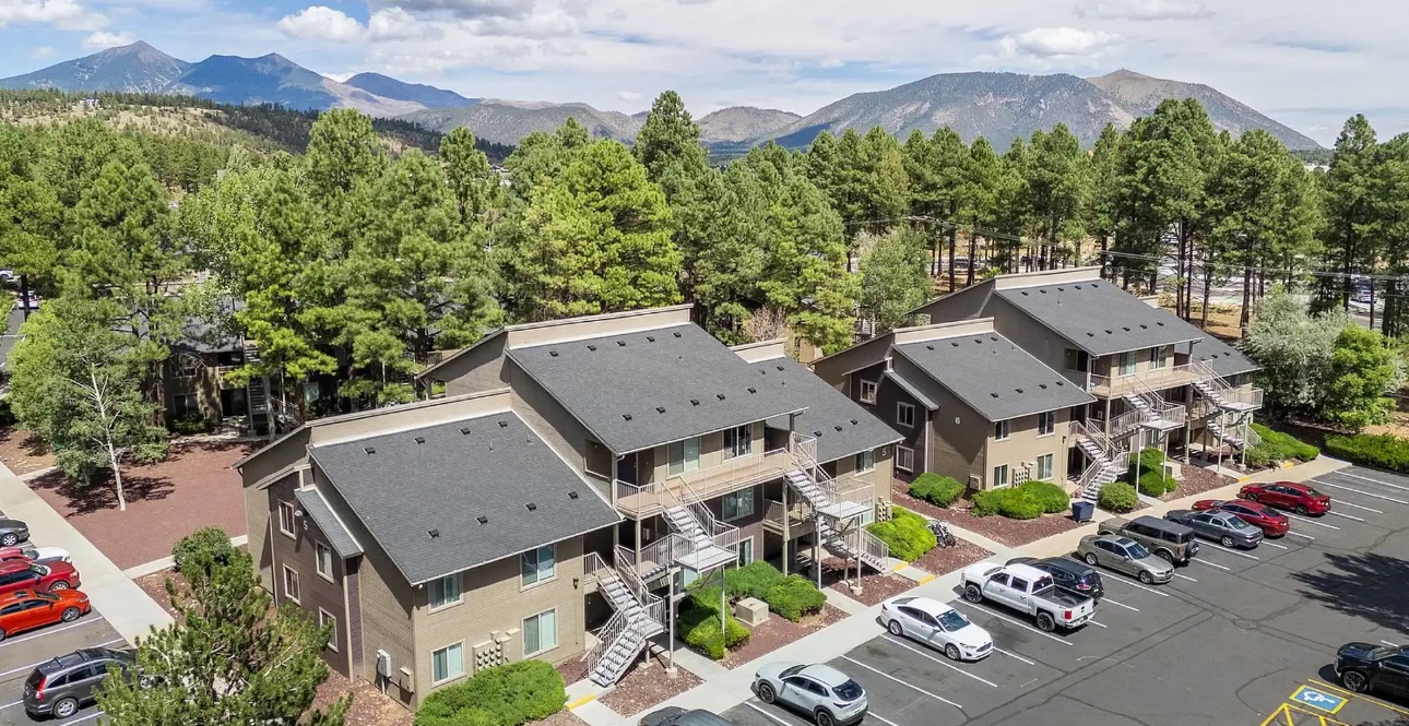 Aerial view of Woodlands Village Apartments Flagstaff, a modern complex nestled among pine trees, with mountains in the background and cars parked in the foreground.
