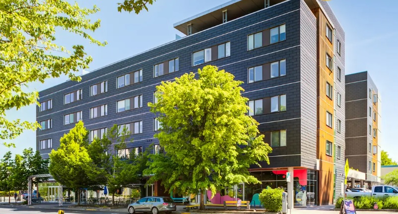 A modern, five-story apartment building—Yugo Eugene Skybox Eugene—with dark siding and yellow accents stands on a sunny street, surrounded by green trees and parked cars.