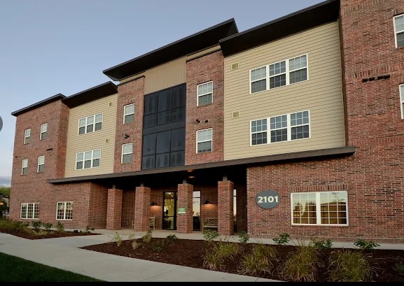 A modern three-story apartment building, Yugo Fort Collins Grove Fort Collins, features red brick and beige siding, large windows, and the number 2101 displayed near the main entrance, all surrounded by attractive landscaping.