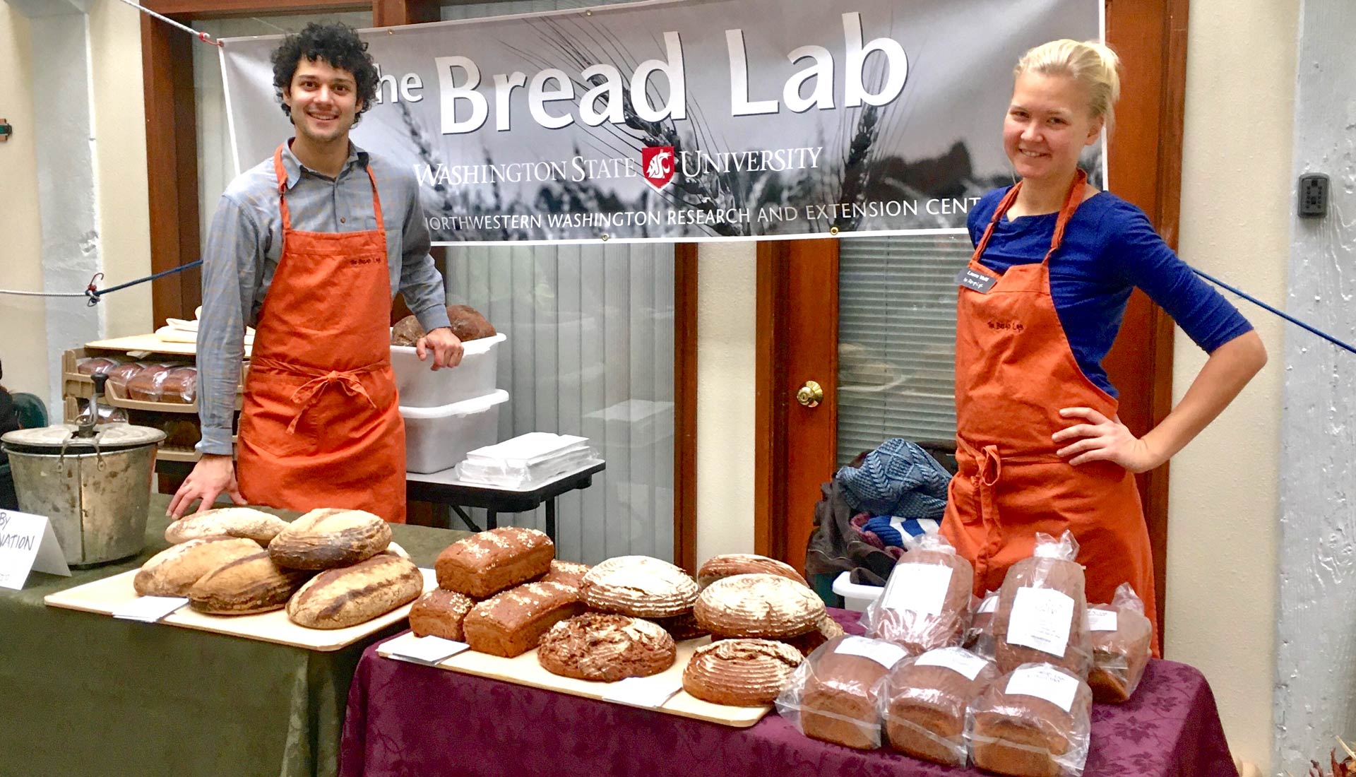 Young man and woman in orange aprons stand at table full of bread.