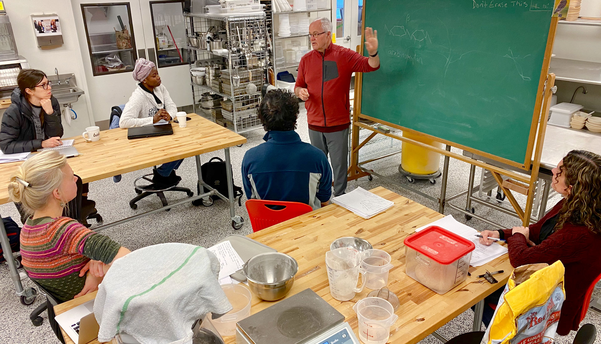 Older man lectures at blackboard to a small group in commercial kitchen