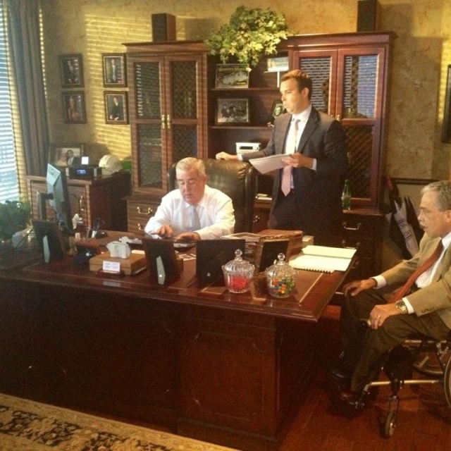 Three men are in a well-furnished office. One sits at a large wooden desk using a computer, another stands with papers, and the third sits in a wheelchair. The mood is professional.