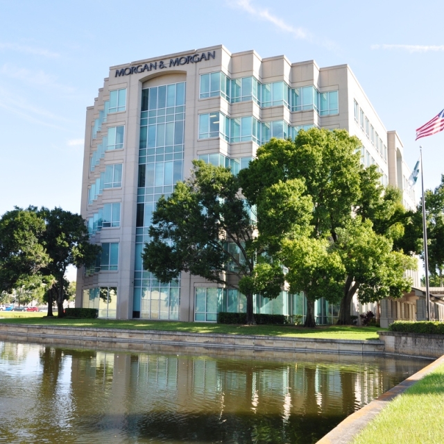 A modern office building with reflective glass windows is situated beside a serene pond. Two large trees and flagpoles with U.S. and company flags stand nearby.