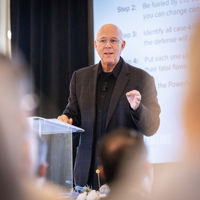 A man in a dark suit speaks confidently at a podium, with a projected presentation behind him.
