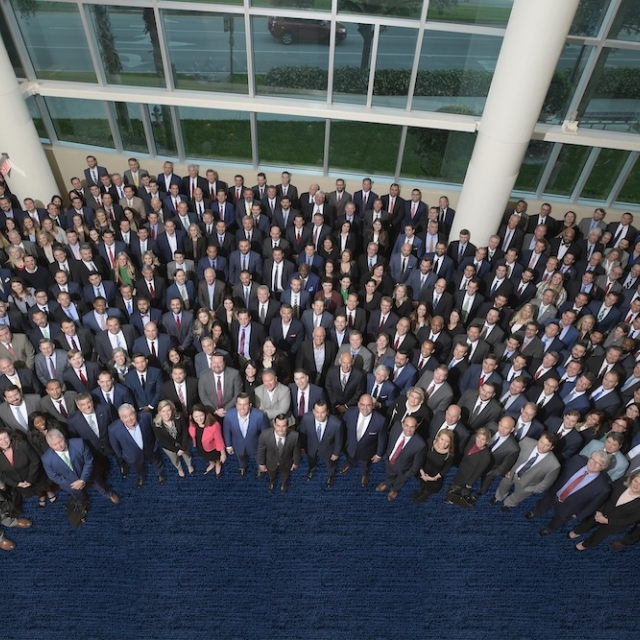 A large group of people in formal attire stands closely together in a bright, spacious indoor area with large windows, conveying a sense of unity and professionalism.