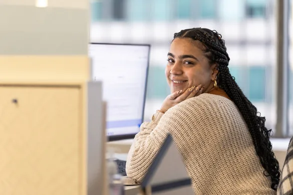 Smiling woman with braided hair and a beige sweater sits at a desk