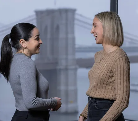 Two people stand in conversation, smiling at each other. They are in front of a large window with the Brooklyn Bridge visible in the background.