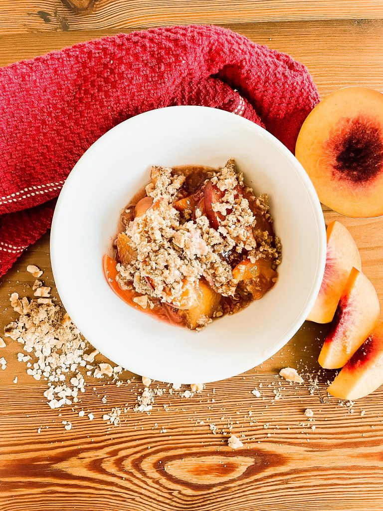 White ceramic bowl with peach crisp inside - red towel, peach slices, and oats scattered on wood table