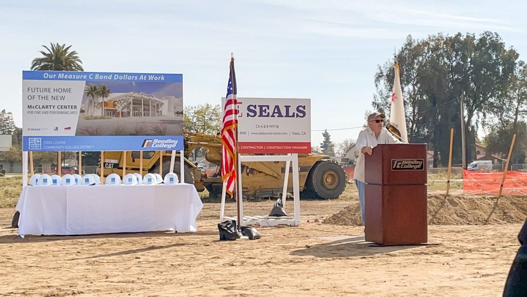 Man standing at podium in a dirt lot. American flag and construction signs at left