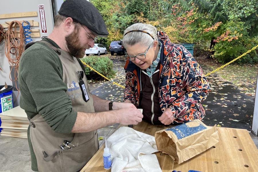 Photo taken of attendees at a recent Fix-It Fair. A man holds a newly repaired soapdish, showing to an attendee near a workbench.
