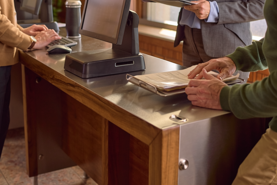A cozy, older government office lobby with wood-paneled walls and a warm, inviting atmosphere. The space includes 3 computer kiosks for public use, with people interacting with the kiosks.