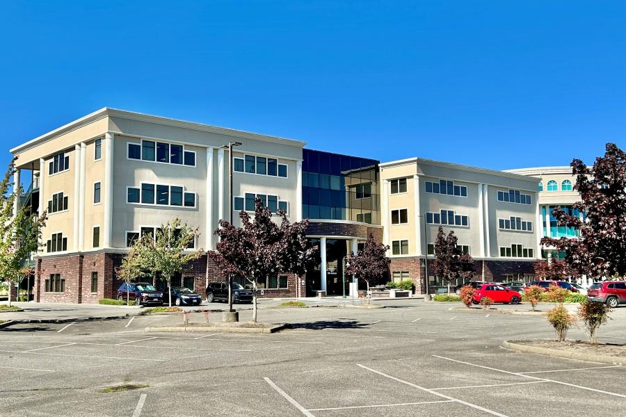 image shows two large modern, multistory office buildings connected by a glassed in breezeway in the center. the view is from the front of the building and its large parking lot on a sunny blue-sky day