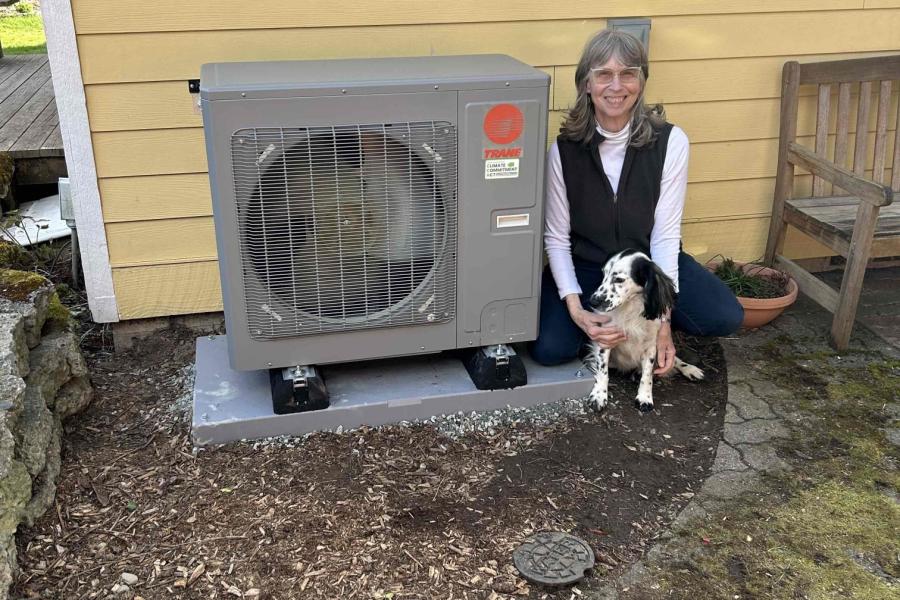 A community member poses with her dog for a photo next to her newly installed Heat Pump unit beside her yellow-painted house as part of the grant-funded Energize Thurston program.