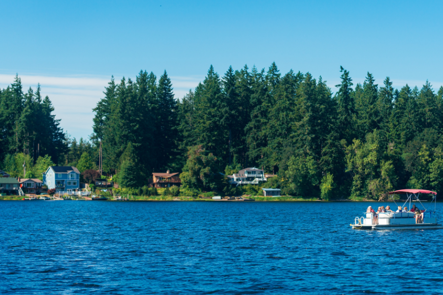 Photo of Offutt Lake in Thurston County on a sunny day. The dark blue lake is edged by tall evergreen trees and a few houses dotting the shoreline. In the mid-distance is a small boat.