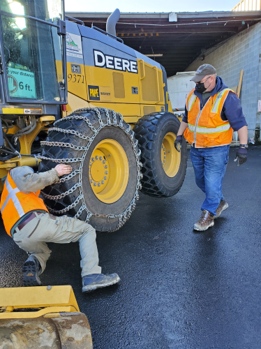 A senior road operations staff member showing a new employee how to put chains on large equipment needed during winter storm operations.