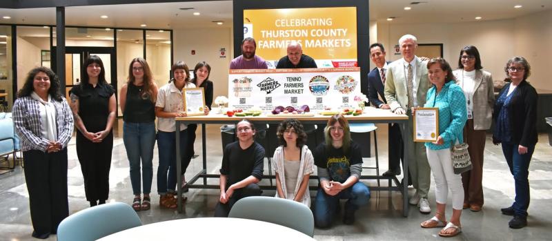Thurston County Commissioners, staff, and members of the community standing next to a sign promoting Thurston County Farmers Markets Week