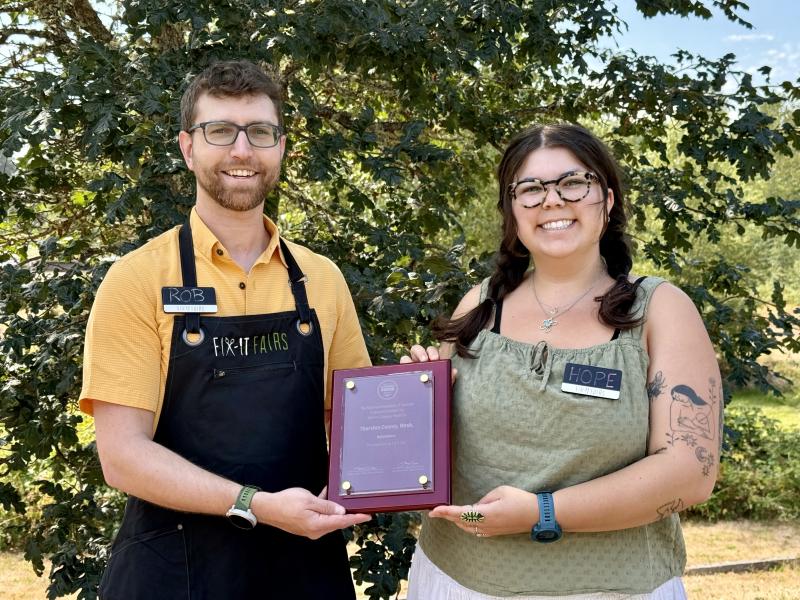 A man in a yellow shirt and black apron and a woman in a green shirt are smiling and holding an award between them.