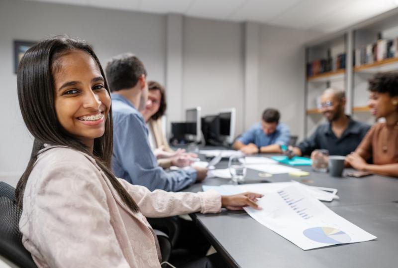 Portrait of a smiling teen sitting in conference room with colleagues discussing in background at office