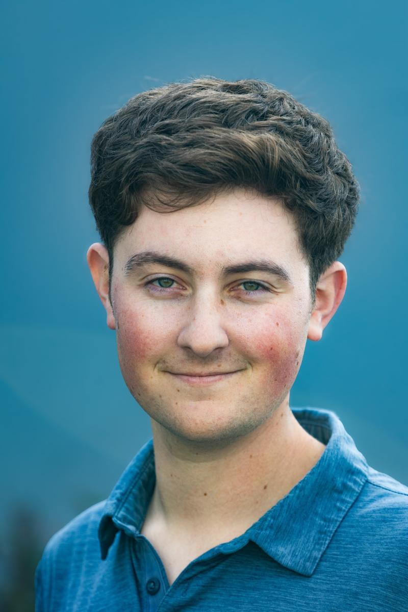 Professional headshot portrait of AJ who has short brown hair and green eyes wearing a blue collared shirt against a blue background.