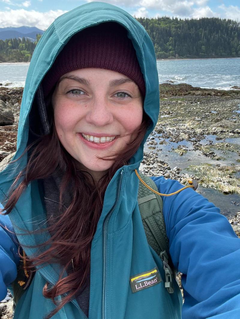 Nym in a teal hooded jacket and burgundy beanie taking a selfie on a rocky shoreline, with forested hills and water in the background.
