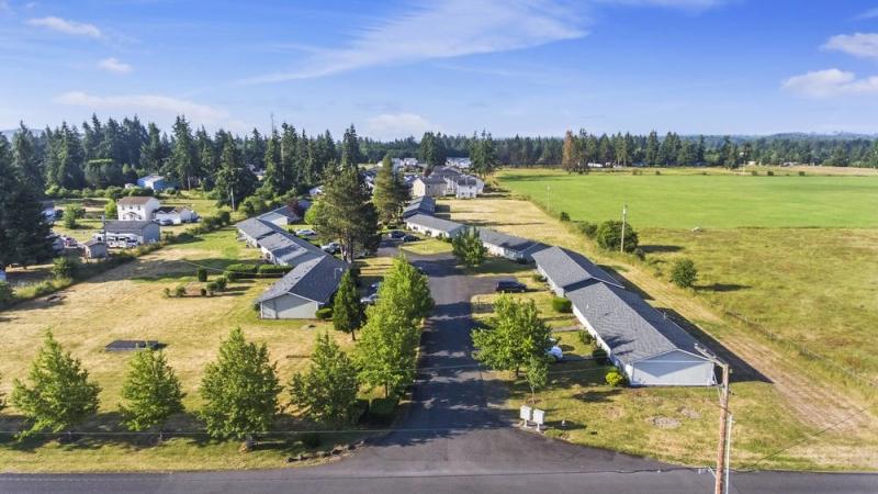 Aerial view of a residential conservation subdevelopment with multiple buildings arranged along a central road, surrounded by evergreen trees and adjacent to a large green field under a blue sky.