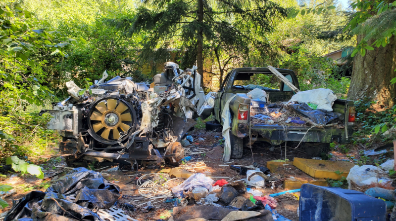 Wrecked truck components and scattered debris piled in a wooded area, illustrating an example of property maintenance and junk vehicle code violations.
