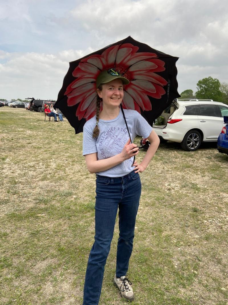 Woman smiling while holding a black umbrella with a large floral design at an outdoor event. She wears a gray t-shirt, jeans, and a green cap, with her hair in a braid. Cars and other attendees visible in the background field.