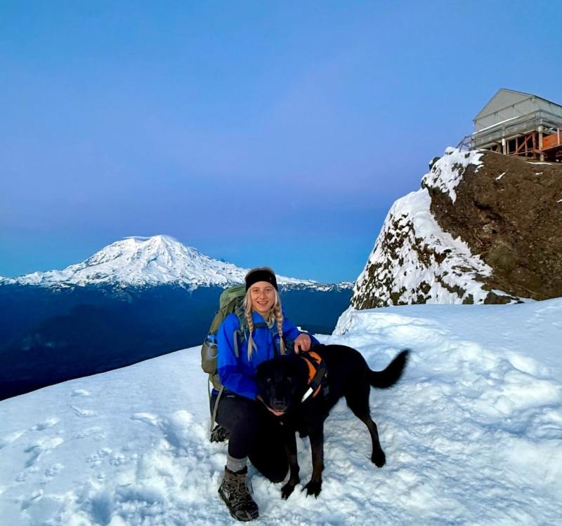 Woman on top of a snowy mountain with a dog. Another mountain in the background.