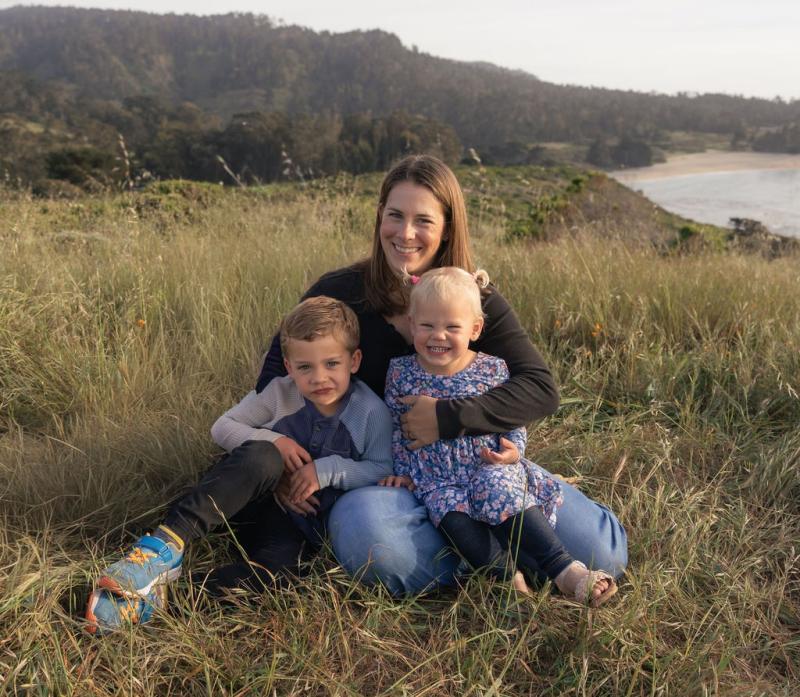 Mother sitting in tall grass on a coastal hillside with her young son in a striped shirt and daughter in a blue floral dress. The family poses together with a scenic backdrop of rolling hills, trees, and beach visible in the distance.