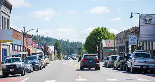 A sunny, wide street view of a small town main street lined with parked cars, trucks, and multi-story storefront buildings on both sides. A dark SUV drives down the center of the two-lane road toward the background. American flags hang from some storefronts under a blue sky with light clouds, and forested hills are visible in the far distance.