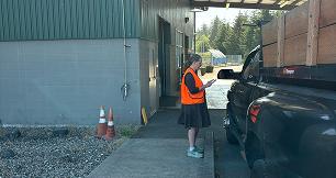 A facility worker wearing a bright orange safety vest stands outside a building next to a black pickup truck. The worker is holding and looking down at a mobile device. The truck has wooden slats extending above its bed. In the background, trees and an open outdoor space are visible under a covered overhang area.