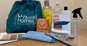 A collection of eco-friendly cleaning supplies arranged on a wooden surface. On the left is a large teal "Healthy Homes" drawstring bag. In front of it sits a small orange box of baking soda, a folded blue microfiber cloth, a scrub brush with a blue handle, and a metal squeegee. In the background are bottles of liquid soap, distilled white vinegar, a white bottle, and an empty plastic spray bottle.