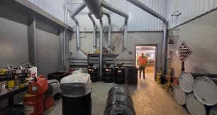 An industrial facility room containing multiple metal barrels and a large ventilation hood system with overhead pipes. On the left are storage shelves, a red gas can, and a large drum filled with white material. Three black barrels sit underneath the vents against the back wall. To the right, a stack of grey barrels rests near a warning sign. A worker wearing a bright orange safety vest stands in the open doorway leading to an adjacent area.