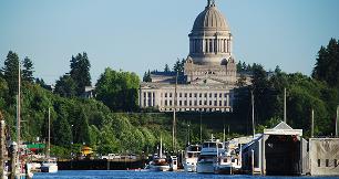 A waterfront view of the Washington State Capitol building sitting on a hill. In the foreground, numerous boats, yachts, and covered docks line a marina. Lush green trees cover the hillside leading up to the large, classic stone capitol building, which features a prominent central dome. The scene is set under a clear blue sky on a bright day.
