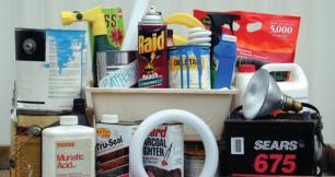 A collection of household hazardous waste items piled together. The items include paint cans dripping with blue paint, a can of bug spray, a bottle of muriatic acid, charcoal lighter fluid, a car battery, fluorescent light bulbs, a floodlight, and various other chemical cleaners, fertilizers, and aerosol cans.